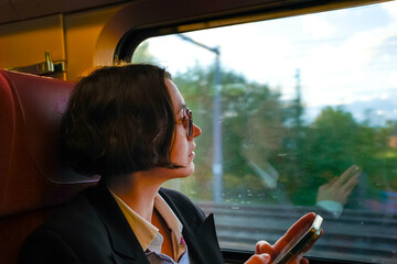 Adult woman wearing glasses looking out the window of a train while holding a smartphone.