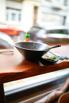 A black empty wok and chopsticks are on a wooden table next to the restaurant's window with a street view.