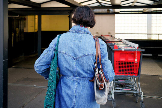 Woman in a denim jacket with a shoulder bag standing near a row of shopping carts.