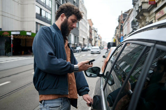 A man in casual attire is using his smartphone while standing next to a parked car in an urban setting, Brussels, Belgium