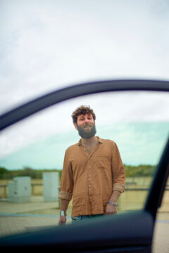 Smiling bearded man standing outside viewed through an open car window with a clear sky in the background.