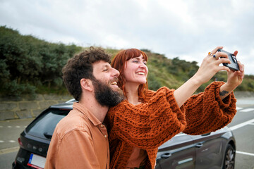 Smiling couple taking a selfie together with a smartphone by their car on a cloudy day.