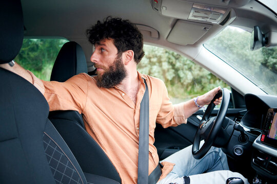 A bearded man driving a car looks back with a puzzled expression.
