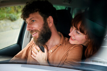 Smiling man with a beard driving a car while a woman with red hair looks at him affectionately.