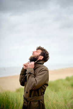 Man in a brown trench coat stands in a grassy field against a cloudy sky, adjusting his collar and looking upwards, Belgium