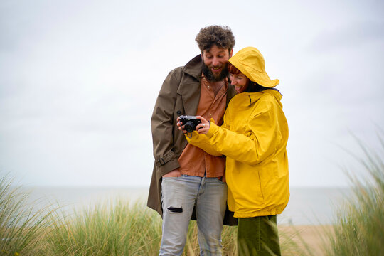 Couple in raincoats reviewing photos on a camera by the beach on a cloudy day, Belgium