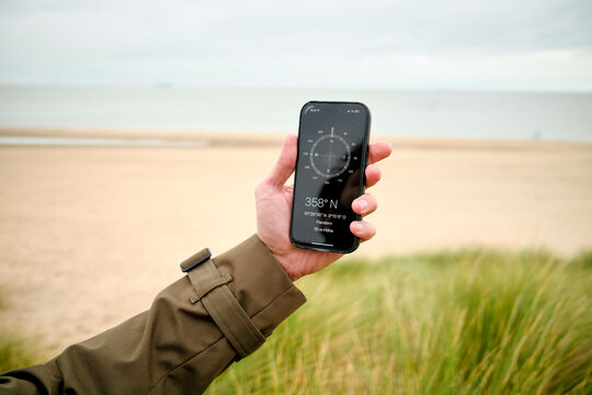 Hand holding a smartphone with a compass app on the screen at a sandy beach under a cloudy sky, Belgium