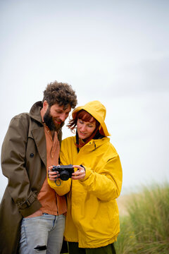 A couple in warm jackets reviews photos on a camera together against a backdrop of tall grass, Belgium