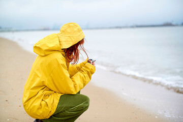 Person in a yellow raincoat crouched by the shore on a cloudy day, holding something, Belgium