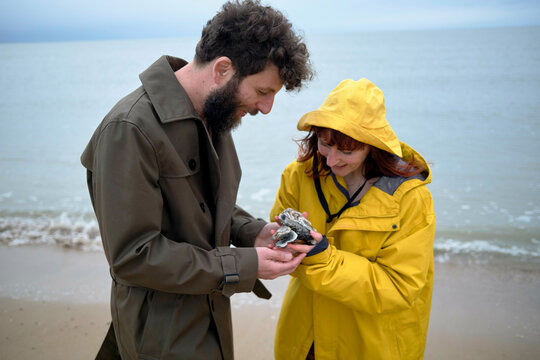 Man and woman in jackets examining sea shells together on a cloudy beach day, Belgium