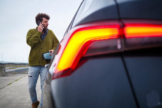 Man talking on a smartphone beside a car with illuminated tail lights on an overcast day, Belgium