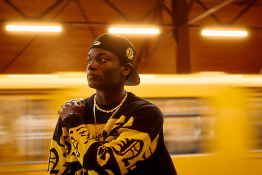 Young man in a black cap and patterned sweater stands pensively against a blurred yellow subway passing by, Berlin, Germany
