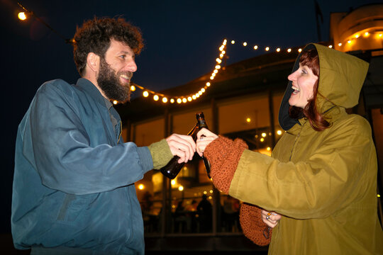 Two adults cheer with bottles under evening lights, Belgium