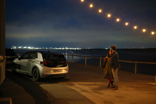 An adult and a child holding hands in a parking lot at twilight with a string of lights above and city lights in the distance, Belgium