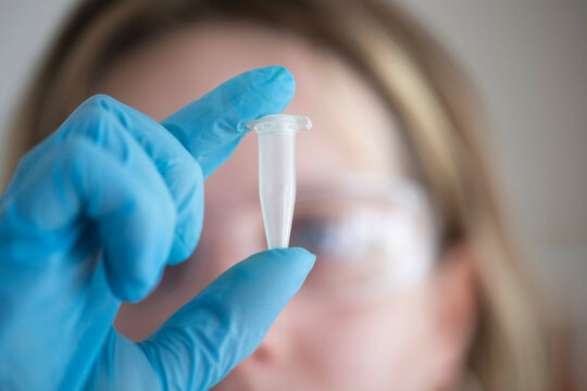 A close-up view of a gloved hand holding a clear test tube in front of a female scientist wearing protective goggles.