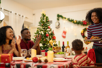 lively holiday dinner scene with a joyful family celebrating Christmas. A woman serves food as others clap, with a Christmas tree, festive decor, and wrapped gifts creating a cozy holiday atmosphere.