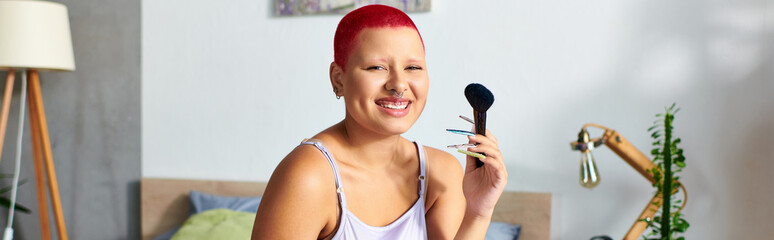 Sitting in her bedroom, a young woman smiles while holding makeup tools, embracing self expression.