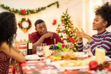 A family enjoys a festive Christmas dinner, sharing food and creating memories. A beautifully decorated Christmas tree and wreath add warmth, while candles and decor enhance the cozy holiday setting.