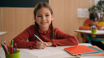 Caucasian clever happy little girl school test study task exam writing notes class lesson learn write copybook kid schoolgirl child pupil studying learning primary education smiling at camera at desk