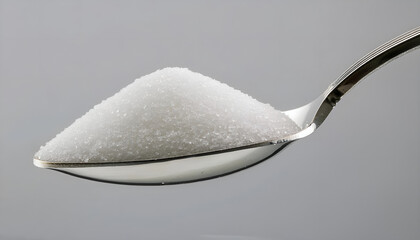 A detailed close-up of white sugar granules on a metal spoon, set against a neutral grey background. Ideal for themes of cooking, sweetness, and food ingredients.