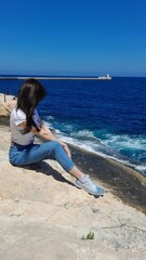 A girl sits on a rock and looks out at the blue sea