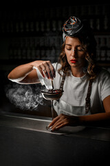 Female bartender holding a wood chip smoldering and blowing smoke around a metal glass