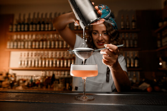 Female bartender pours the last drops of a foamy drink onto a strainer, through which she strains the cocktail into a glass