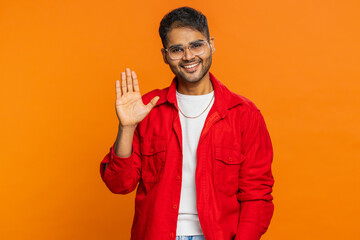 Young Indian man smiling friendly at camera, waving hands gesturing hello greeting or goodbye welcoming with invitation hospitable expression. Arabian Hindu guy isolated on orange background indoors © Andrii Iemelianenko
