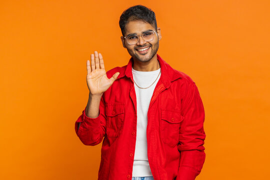 Young Indian man smiling friendly at camera, waving hands gesturing hello greeting or goodbye welcoming with invitation hospitable expression. Arabian Hindu guy isolated on orange background indoors