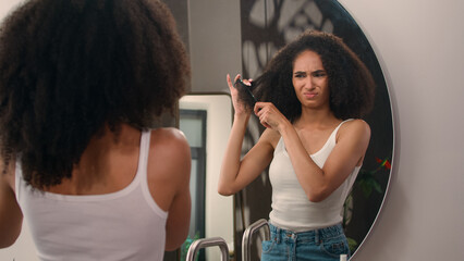African American woman with curls suffer with haircare problem looking at mirror reflection young...