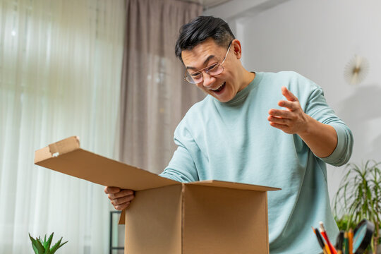Happy Asian mature man unpacking delivery parcel at home. Smiling satisfied Japanese middle-aged guy shopper online shop customer opening cardboard box receive purchase gift by fast postal shipping