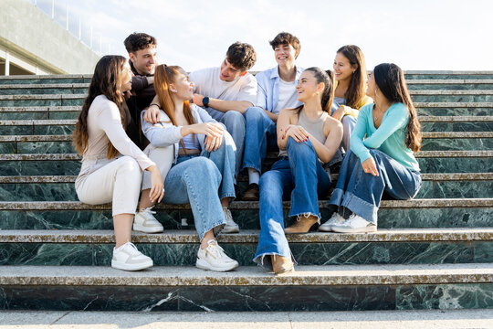 Medium shot photo of a group of 8 young people of different ethnicities between 18 and 25 years old having fun in a city.The millennials are sitting on some steps while chatting.