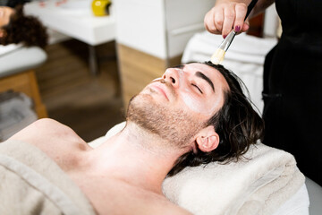 A handsome bearded man in his 30s and 40s is inside a spa.The male is lying on a table while undergoing a facial massage with a brush.Concept of massages with brushes and oil.