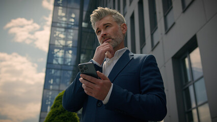 Mature senior employer middle-aged Caucasian entrepreneur outdoors old business man looking at mobile phone thinking pensive ponder idea solution think male businessman in city browsing smartphone
