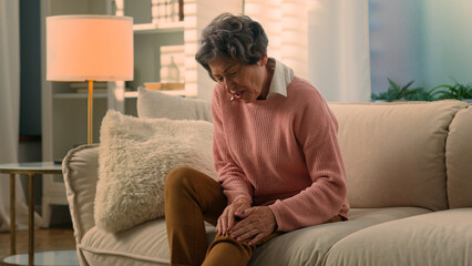 Caucasian old woman tying to stand up from home couch feel sudden pain in knee unhealthy suffering...