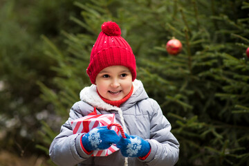 Happy little smiling girl in red cap with Christmas gift box standing near by spruce. Holiday celebrate concept.