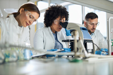 Obraz premium Mid adult female scientist looking through a microscope while working in laboratory with her colleagues.
