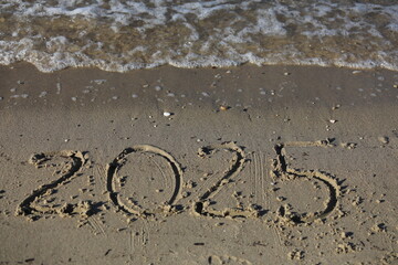 New Year 2025, lettering on the beach with wave and clear blue sea. Numbers 2025 year on the sea shore, message handwritten in the golden sand on beautiful beach background. New Years concept.