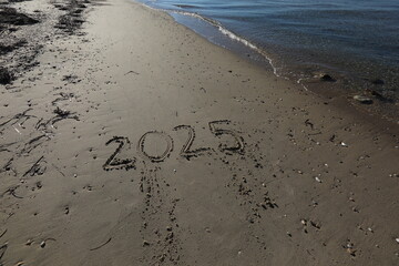 New Year 2025, lettering on the beach with wave and clear blue sea. Numbers 2025 year on the sea shore, message handwritten in the golden sand on beautiful beach background. New Years concept.