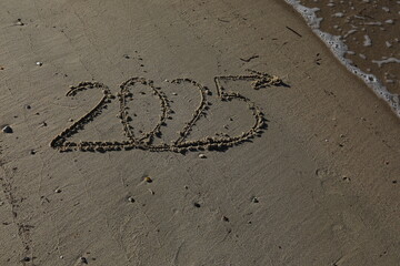 New Year 2025, lettering on the beach with wave and clear blue sea. Numbers 2025 year on the sea shore, message handwritten in the golden sand on beautiful beach background. New Years concept.