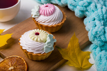 Two tartlets topped with whipped cream, decorated with colorful icing, on wooden board with dried orange slices, autumn leaves, with warm cup of tea and cozy blanket. Ideal for seasonal dessert themes