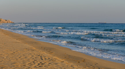 Obraz premium Waves gently lap at the sandy shore under a tranquil sky during sunset near an ocean coastline.