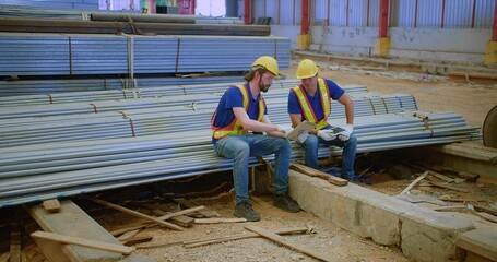 Two construction workers in safety gear review plans on a site holding digital tablet sitting on steel beams, showcasing teamwork and industrial progress.