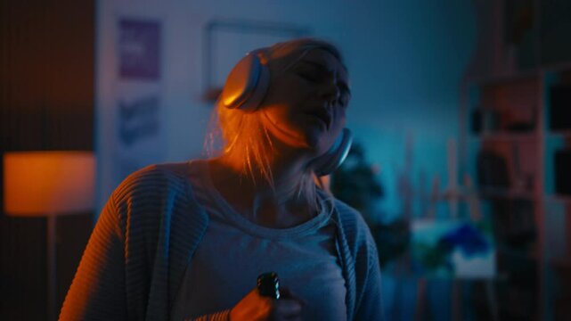 Woman listening to music in headset, dancing and drinking wine alone at home