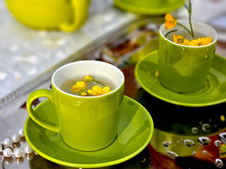 Stylish outdoor tea setting with vibrant green ceramic cups and saucers, accented by delicate yellow flowers floating in water. An elegant and inviting scene on a reflective silver tray