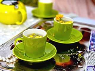 Stylish outdoor tea setting with vibrant green ceramic cups and saucers, accented by delicate yellow flowers floating in water. An elegant and inviting scene on a reflective silver tray