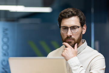 Close-up photo of a thoughtful young businessman sitting in the office at the workplace and looking at the laptop screen while rubbing his beard with his hand