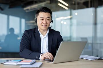 A smiling young Asian man is sitting in the office at a desk wearing a headset and working on a laptop