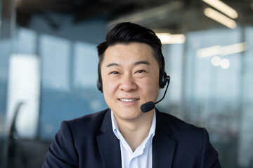 Close-up portrait of a young Asian man in a suit and headset in the office, sitting in front of the...