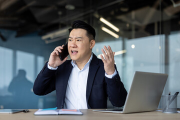 An angry young Asian male businessman is in the office and emotionally talking on the phone while...